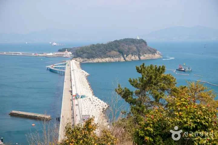 Vista aérea desde el Parque Jasan mostrando la pasarela hacia la isla de Odongdo en Yeosu, rodeada por el mar azul y bosques de camelias de invierno.