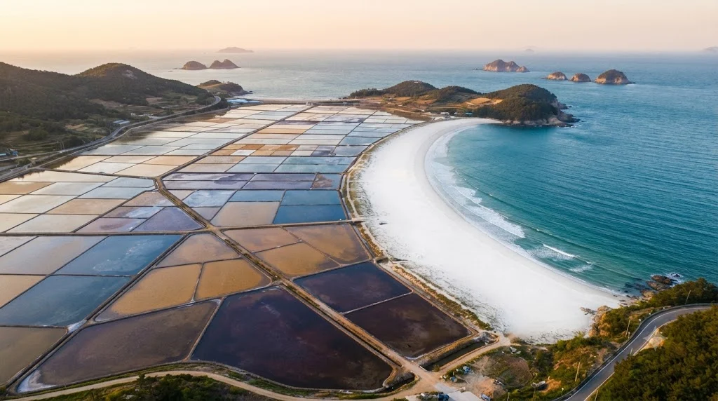 Aerial view of Bigeumdo island in Shinan, South Korea, showcasing geometric solar salt evaporation ponds adjacent to a pristine crescent beach and the Yellow Sea at sunset.